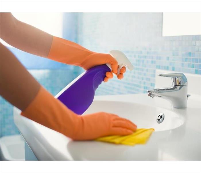Orange gloved hands holding a purple spray bottle and yellow rag wiping a sink